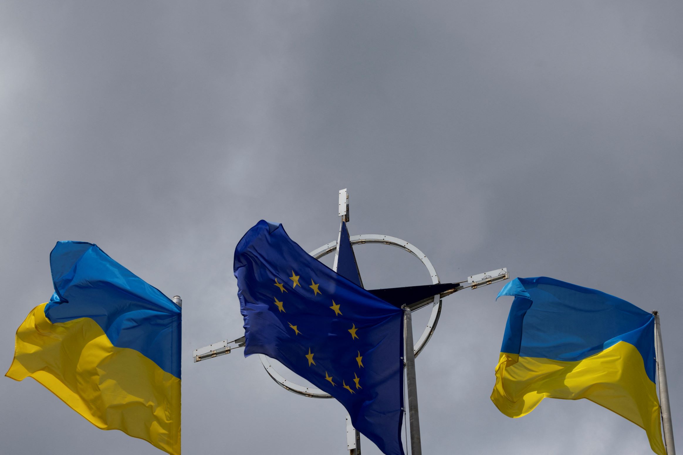 Two national flags of Ukraine flank the European Union flag in the foreground to the NATO emblem with storm clouds in the background in central Kyiv, Ukraine on July 11, 2023. REUTERS/Valentyn Ogirenko